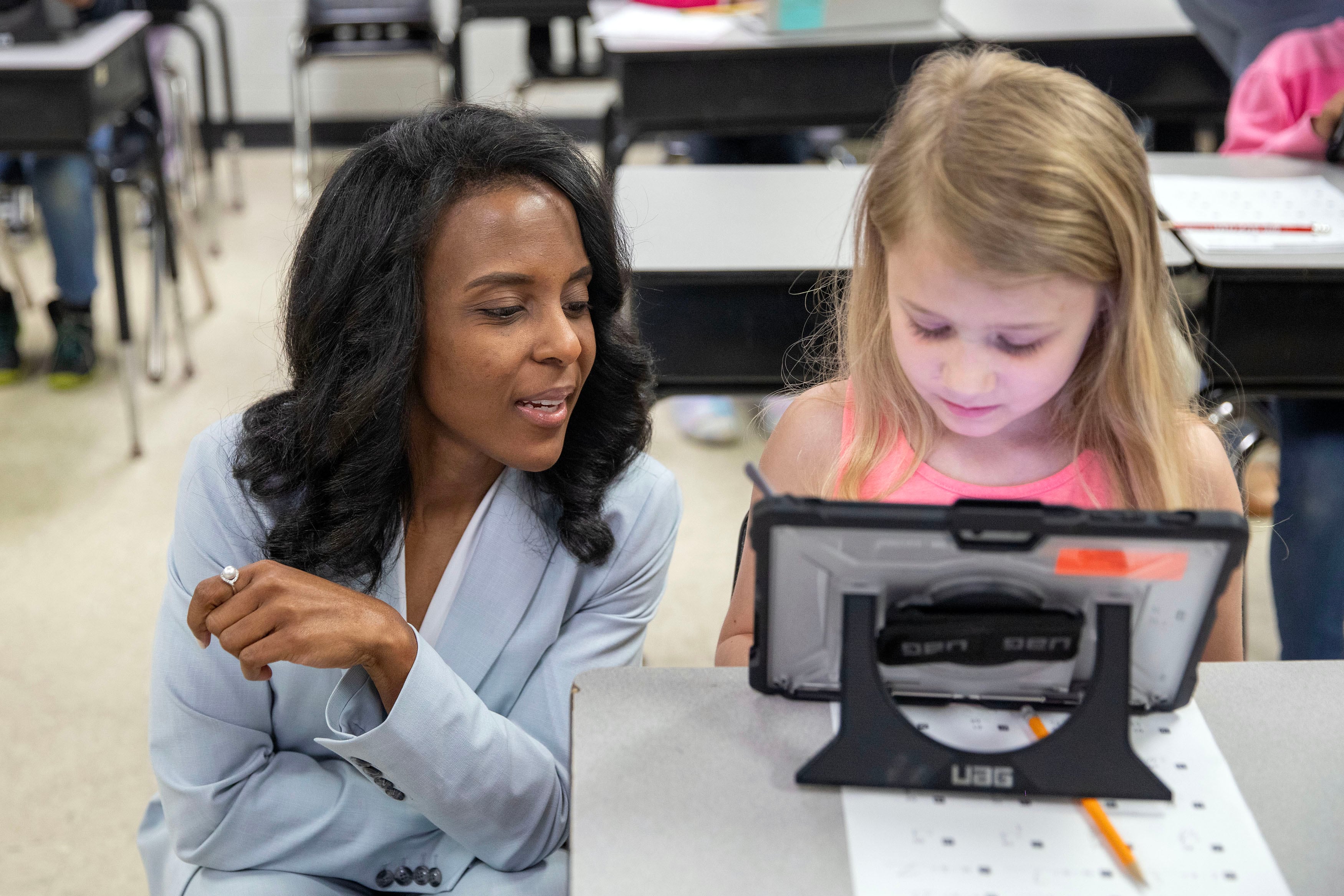 A woman in a blue suit kneels next to a young student while she works on her tablet in a classroom.