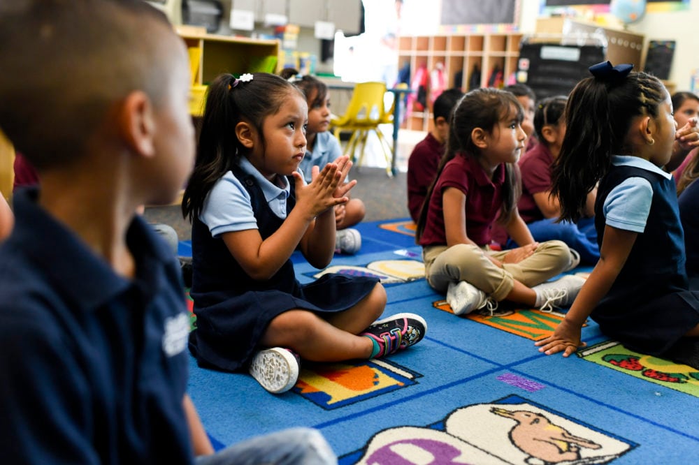 A group of young students sit on a colorful rug in a classroom.