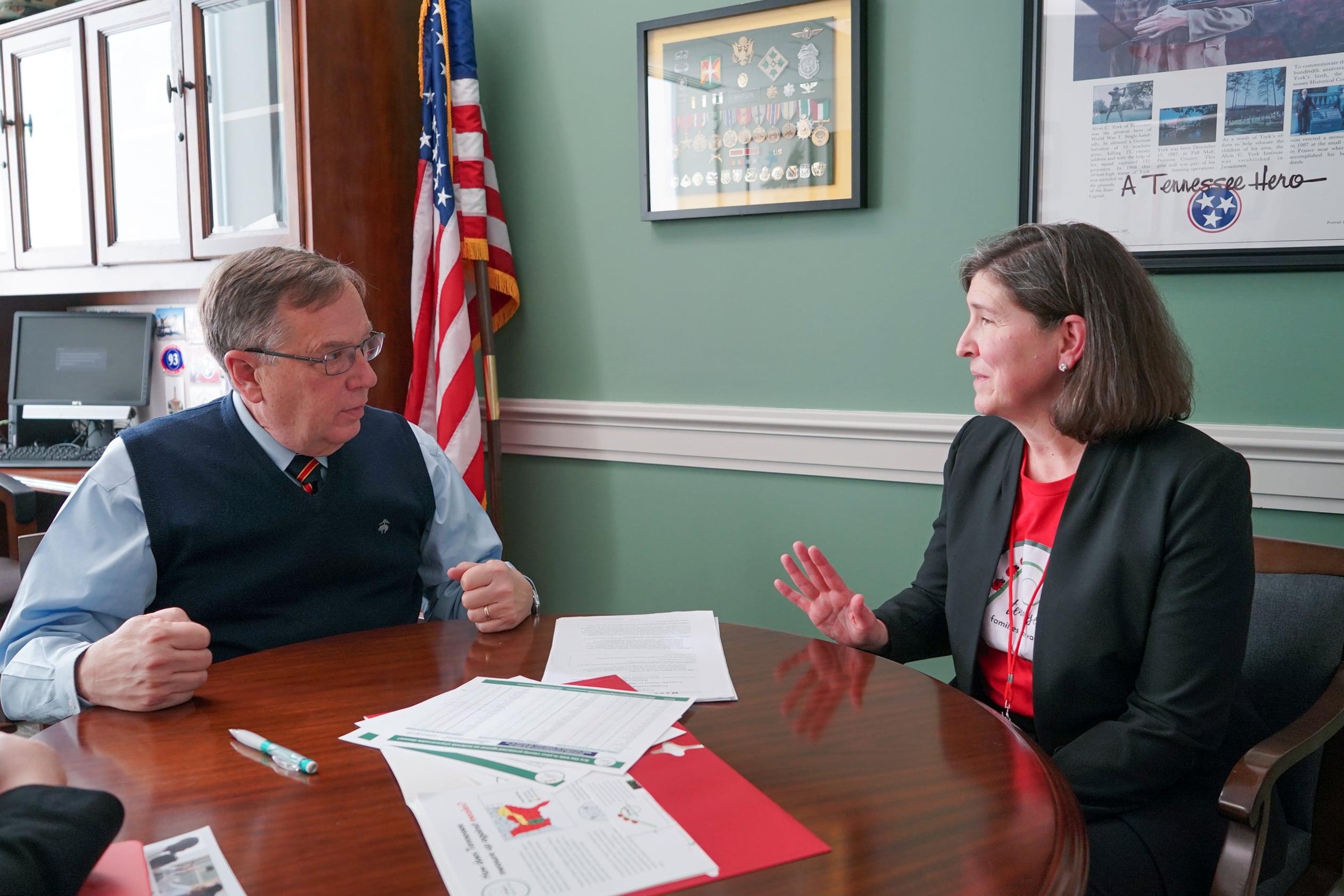 Two people wearing business clothes sit next to each other at a round, wooden table with pictures and a flag in the background.
