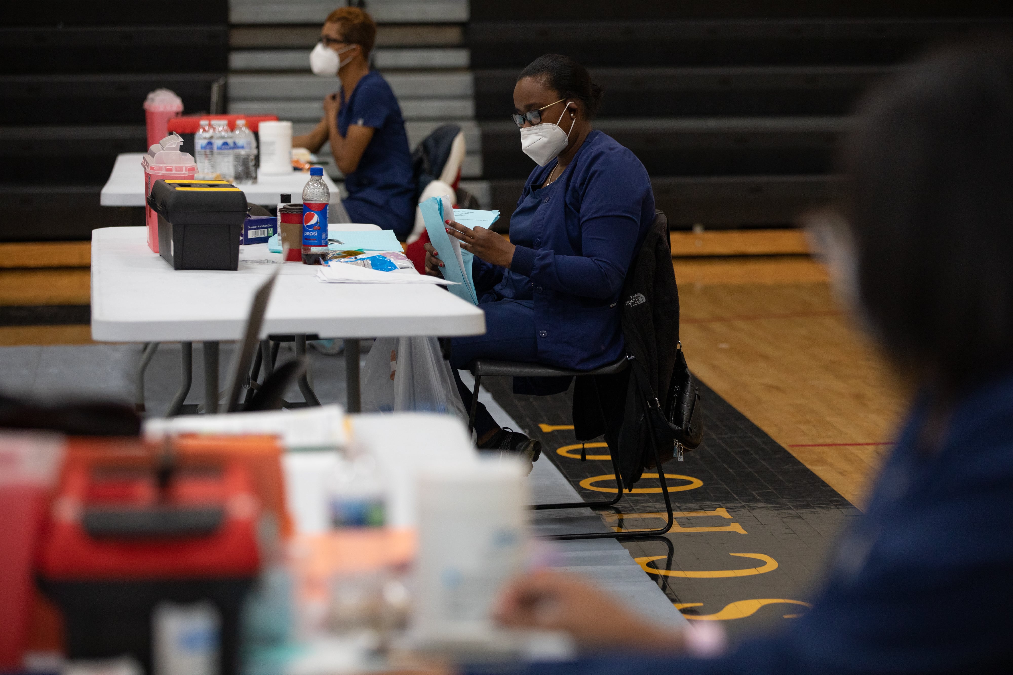Three nurses sit at a row of tables set up inside of a school gymnasium. The person in the middle is is wearing a mask while they flip through a series of packets.