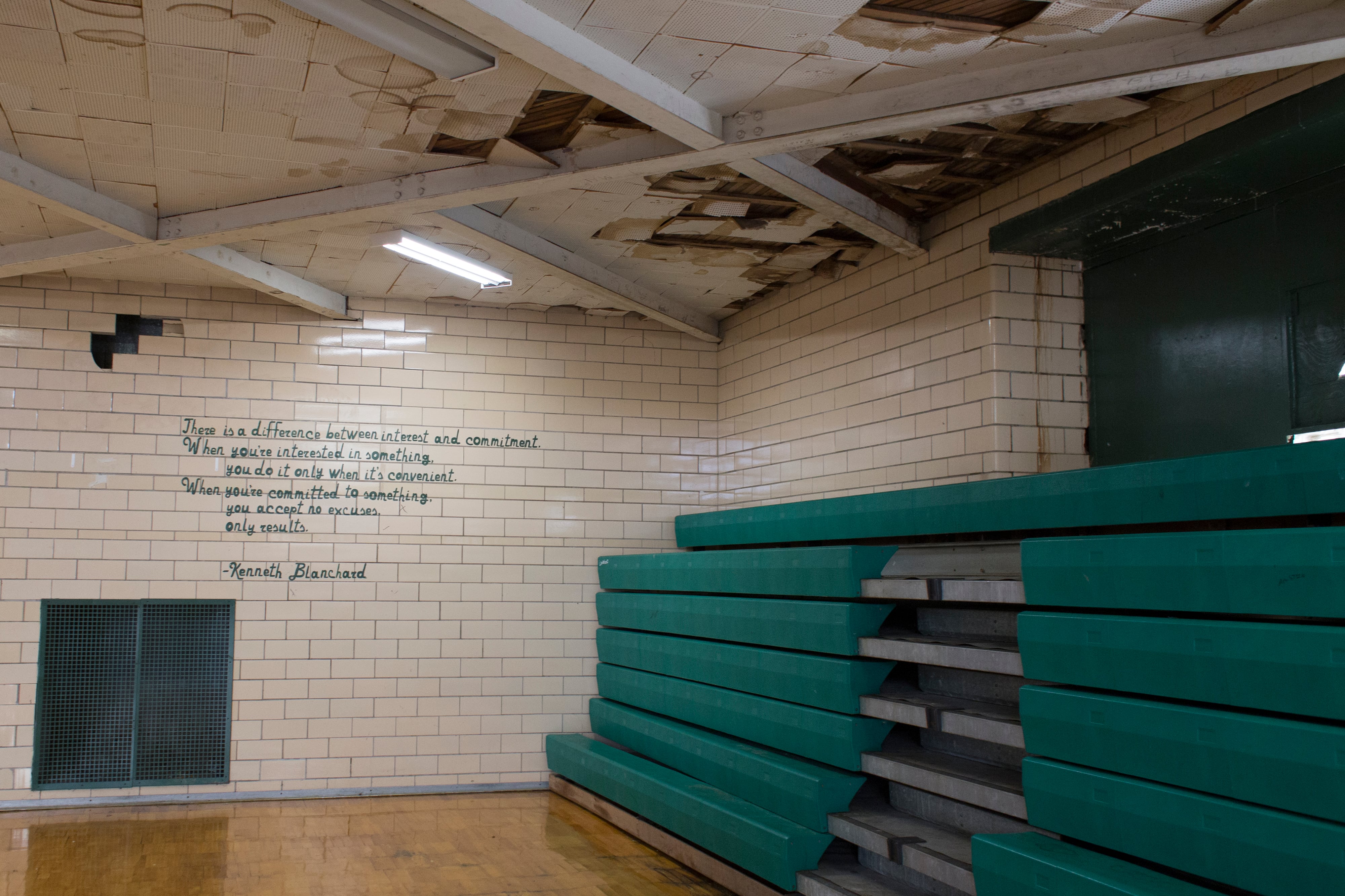 The ceiling tiles are missing in a corner of a high school gym with green bleachers on one side and a brick wall and black writing on the other.