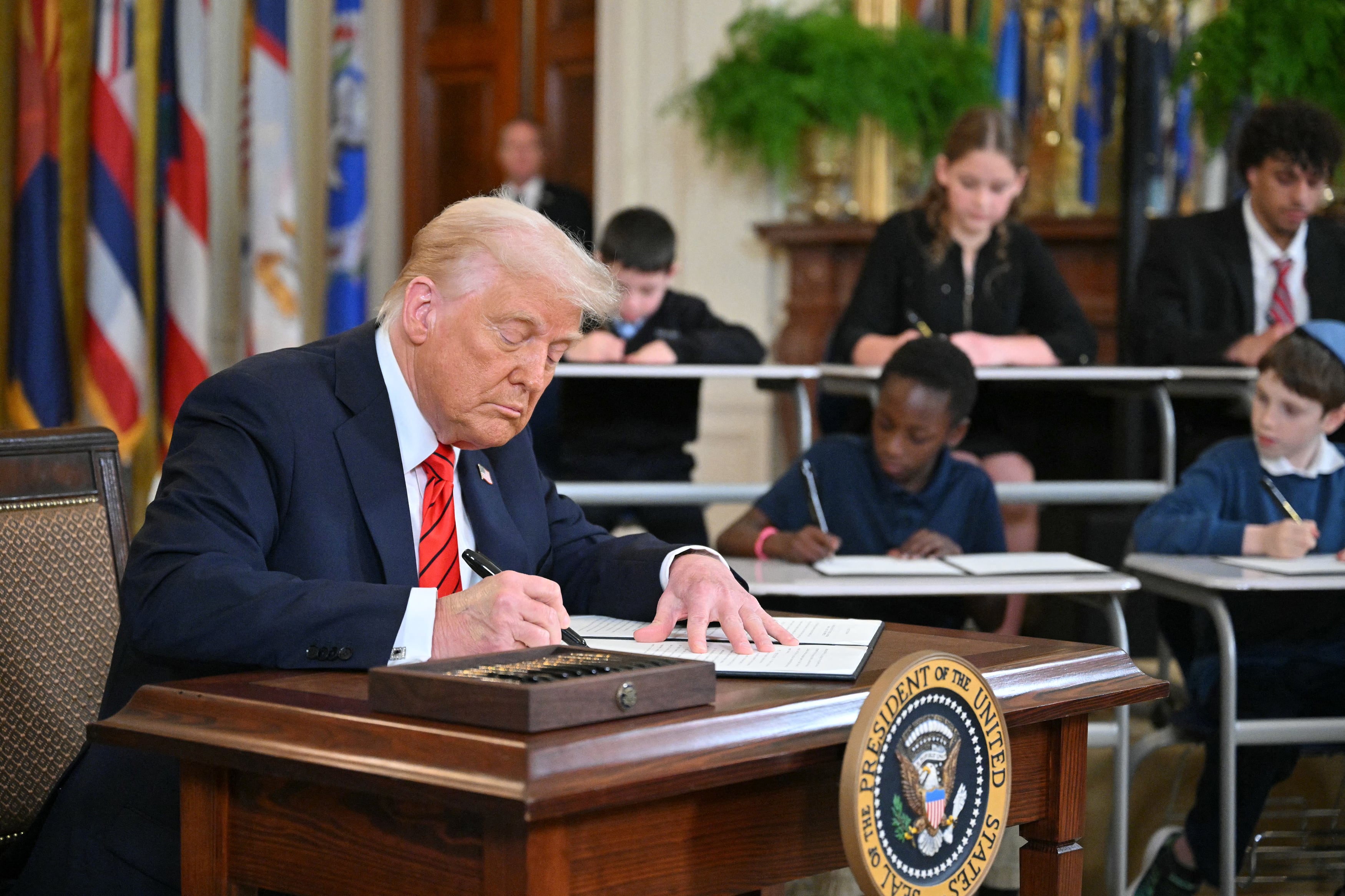 A white man with white hair and wearing a dark suit and a red tie sits at a wooden table signing a piece of paper with two rows of young children sit at desks in the background all also signing papers.