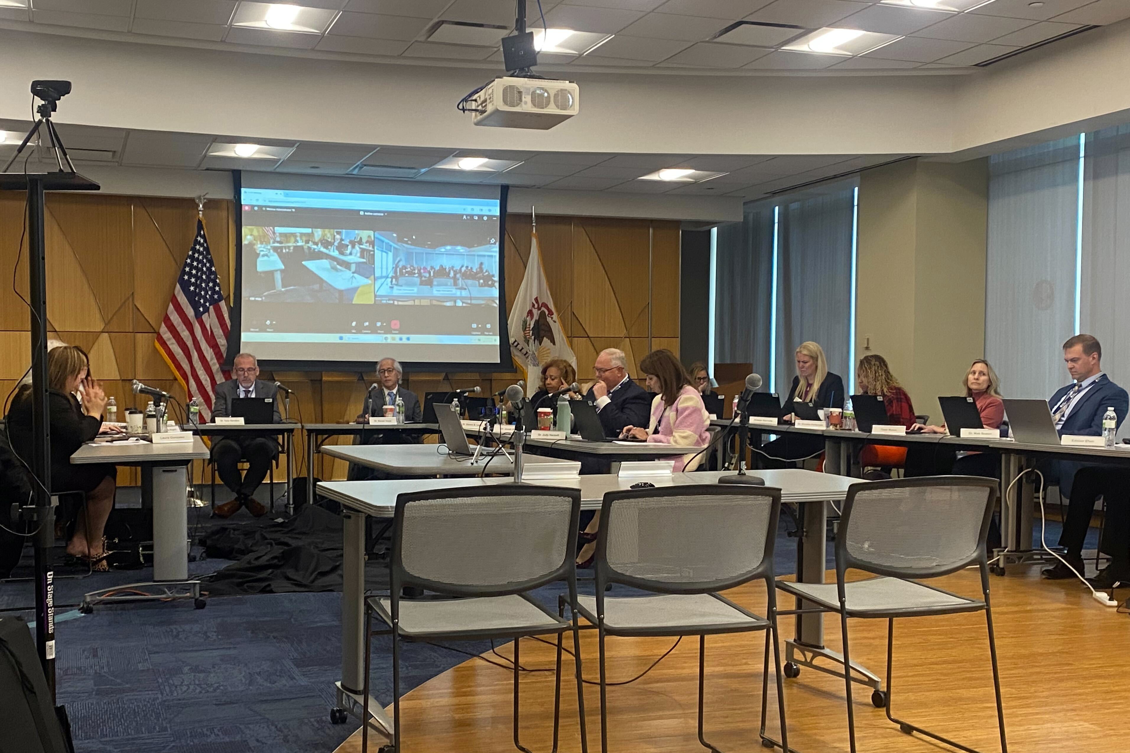 A photograph of a group of adults in business clothes sit around tables in a meeting room.