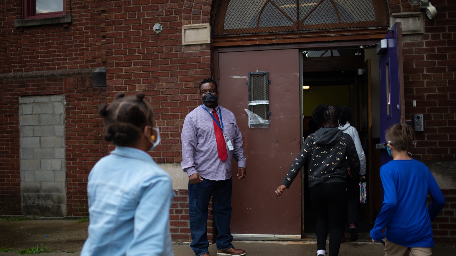 A teacher watches as students make their way into the brick facade of their school.