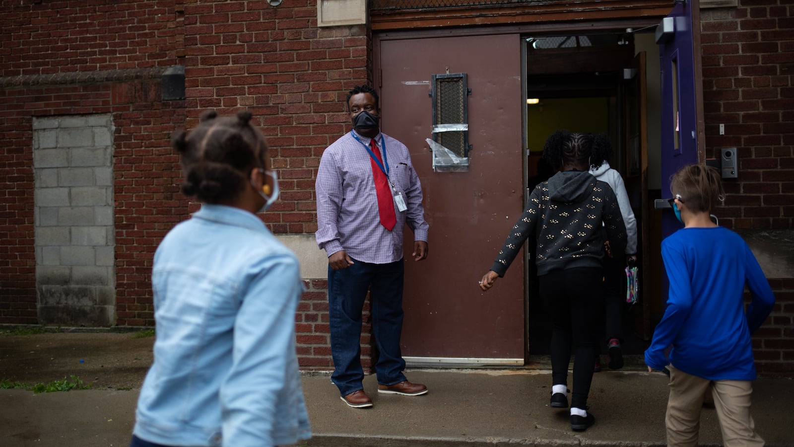 A teacher watches as students make their way into the brick facade of their school.