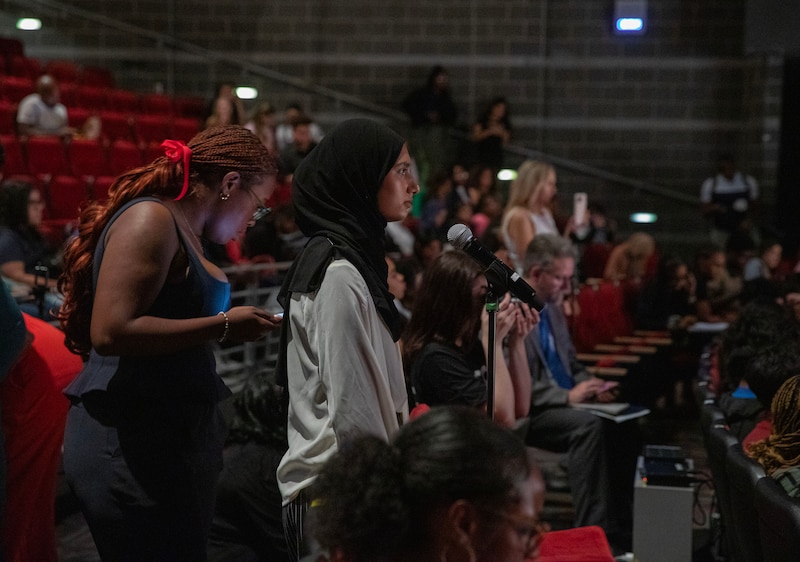 A high school student stands behind a microphone and in front of a group of students in a dark school auditorium.