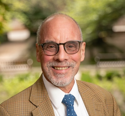 Headshot of a man wearing glasses, a beige tweed blazer and a blue tie