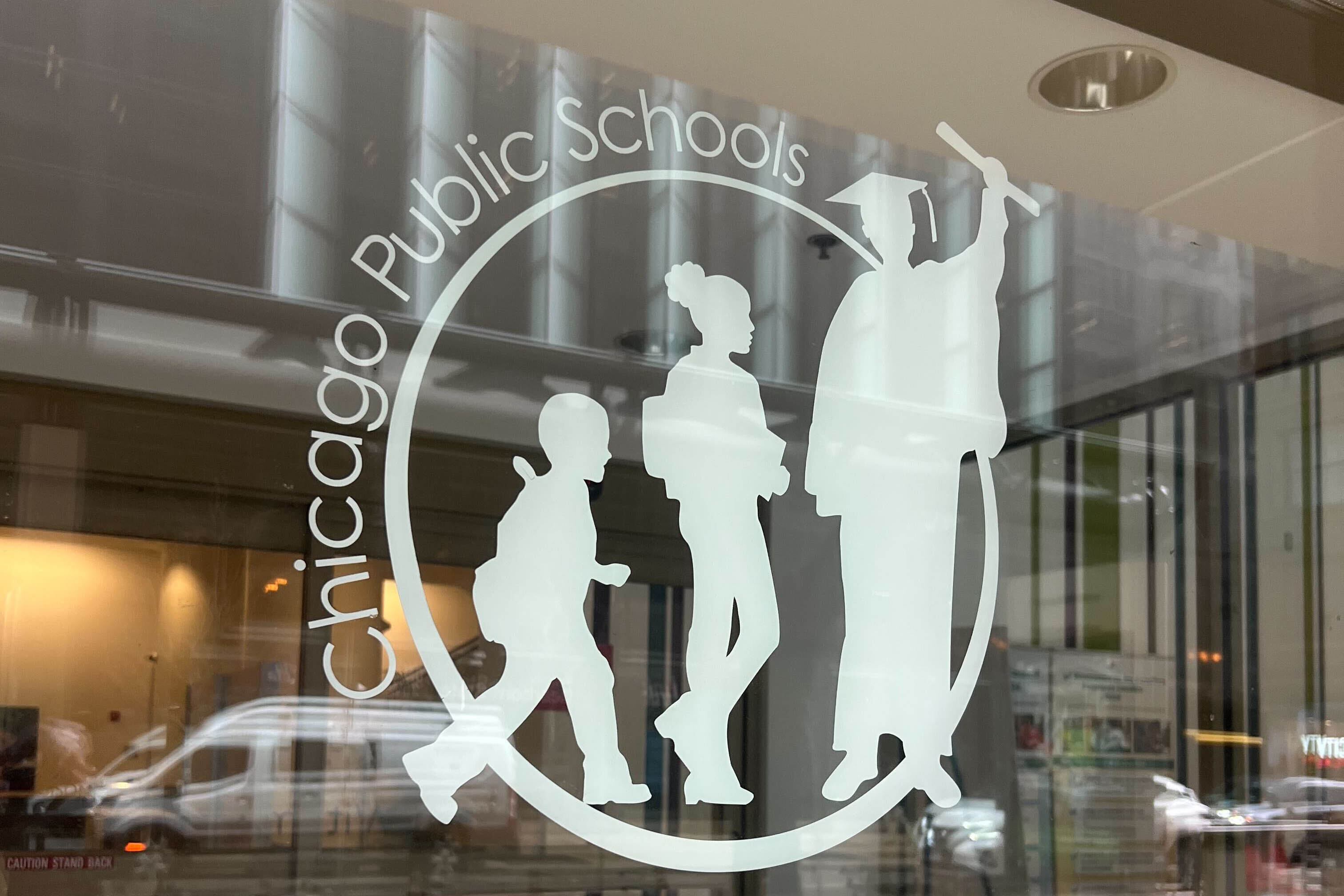 The Chicago Public Schools logo on the side of a glass wall with reflection of cars and people passing in the background.