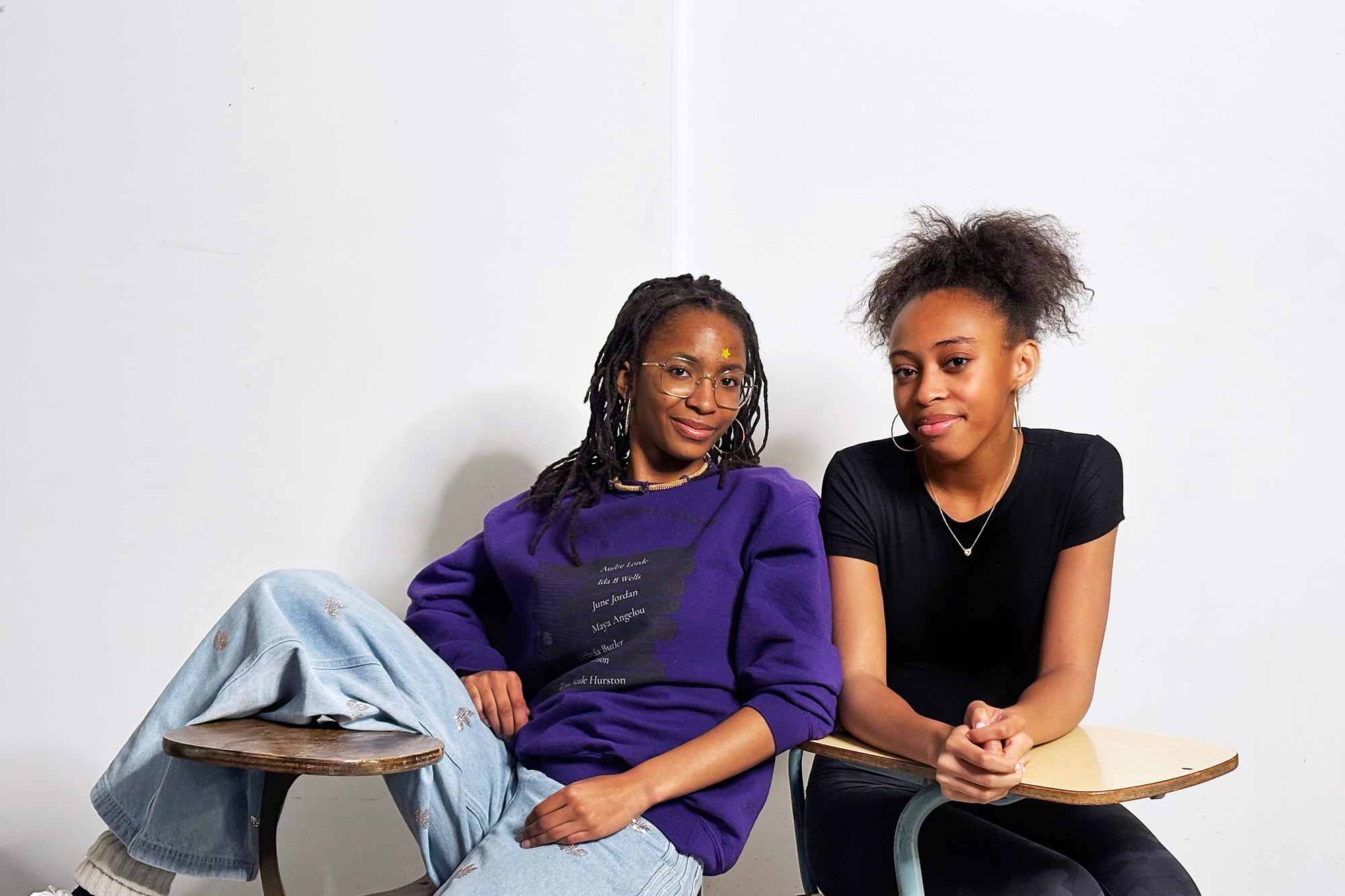 Two Black high school students sit next to each other on school desks in front of a white wall and pose for a portrait. The student on the left has long dark hair and is wearing a purple sweater and blue jeans and the student on the right has dark hair in a ponytail and wearing a black t-shirt and dark jeans.