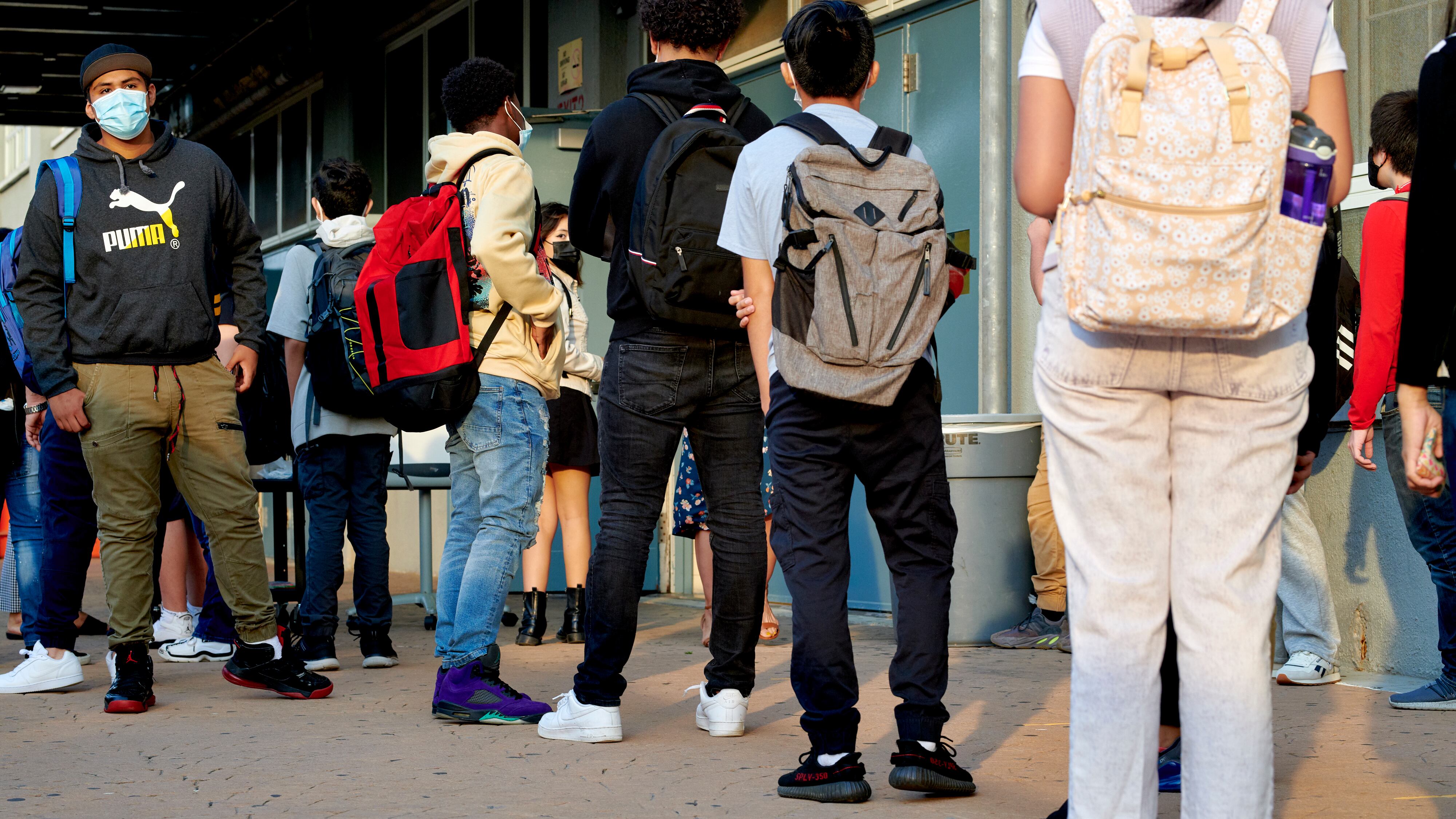 Students, wearing masks and backpacks, wait in line outside a school. Most are visible from the back only.