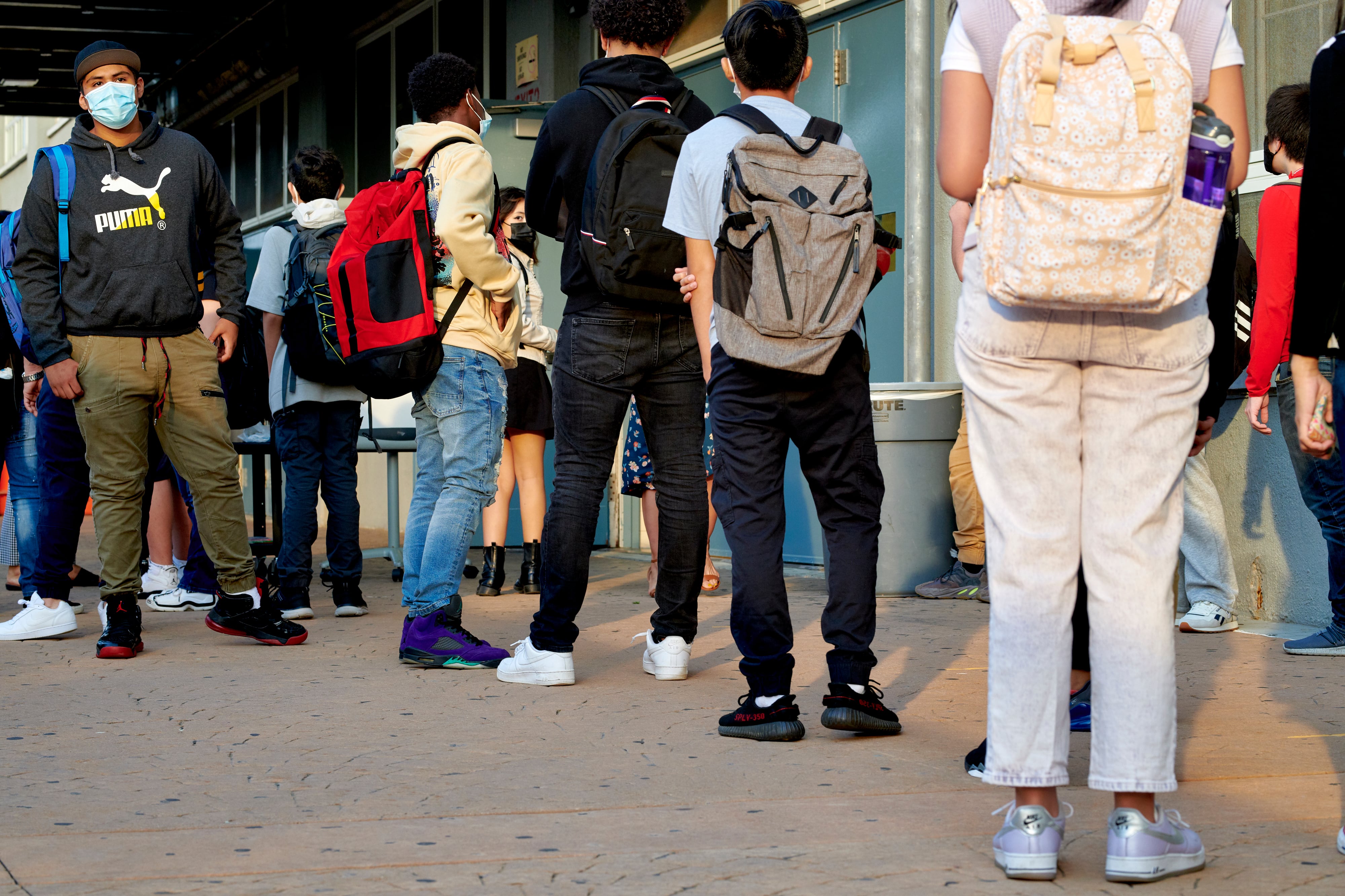 Students, wearing masks and backpacks, wait in line outside a school. Most are visible from the back only.