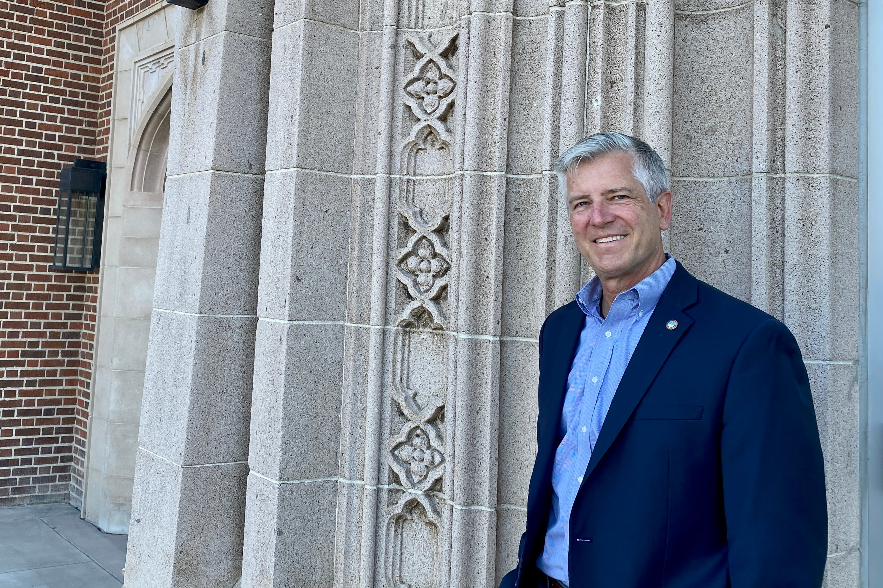 A man in a suit jacket and button down shirt stands in front of a school smiling at the camera