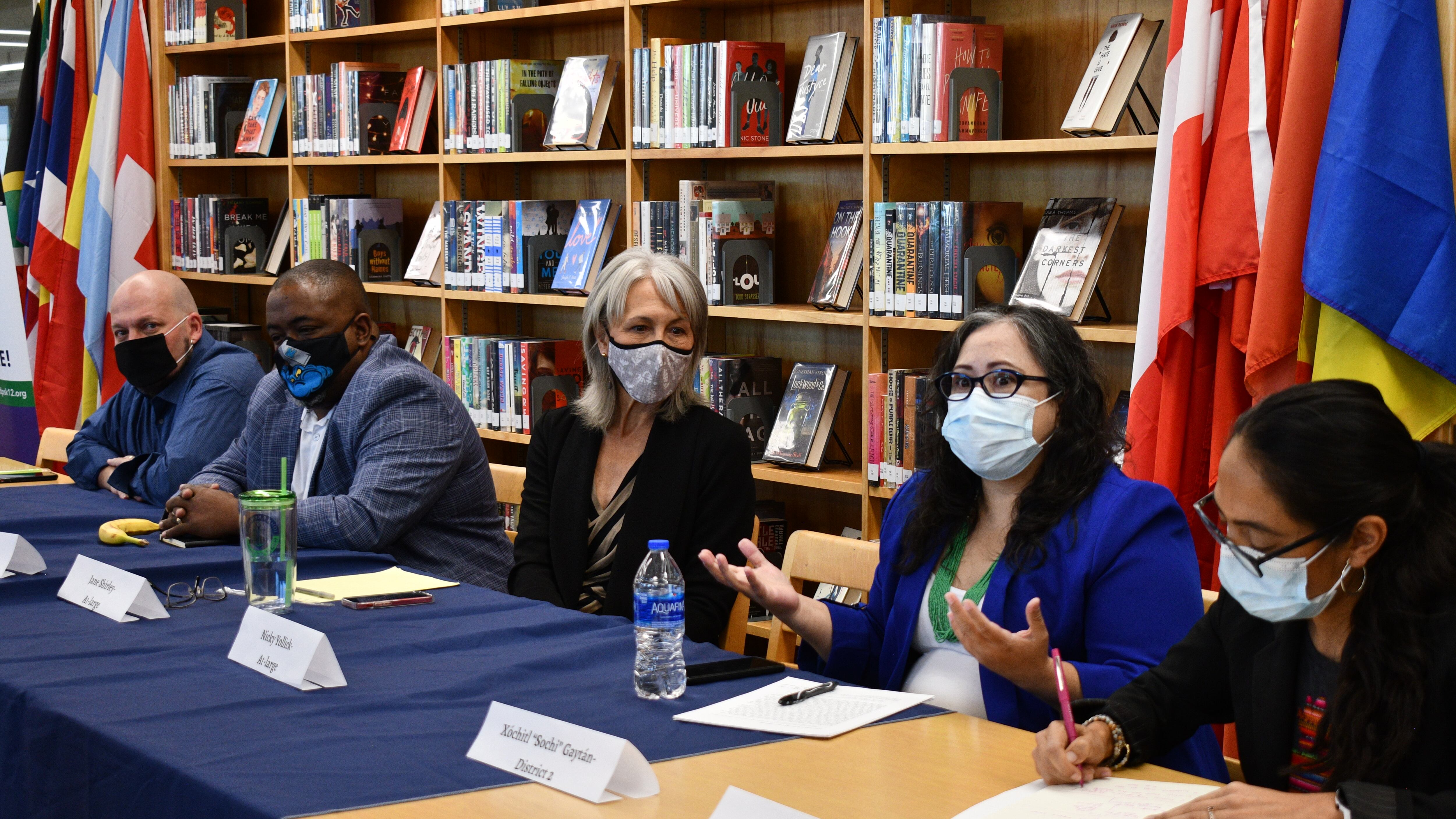 Five Denver school board candidates sit at a table in a high school library during a candidate forum. The candidates are wearing masks.