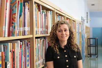 A photograph of a white woman with curly hair and wearing a black blouse poses for a portrait in a school library.