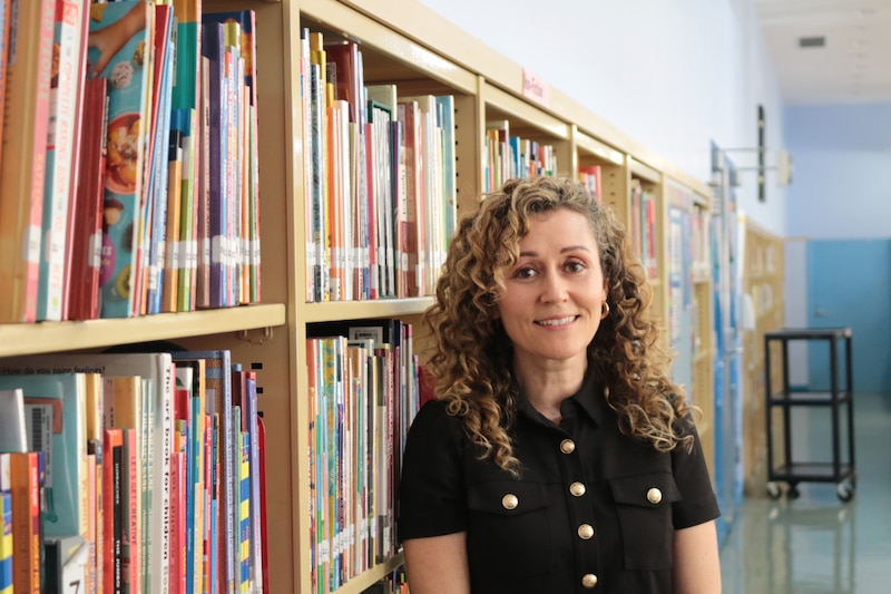 A photograph of a white woman with curly hair and wearing a black blouse poses for a portrait in a school library.