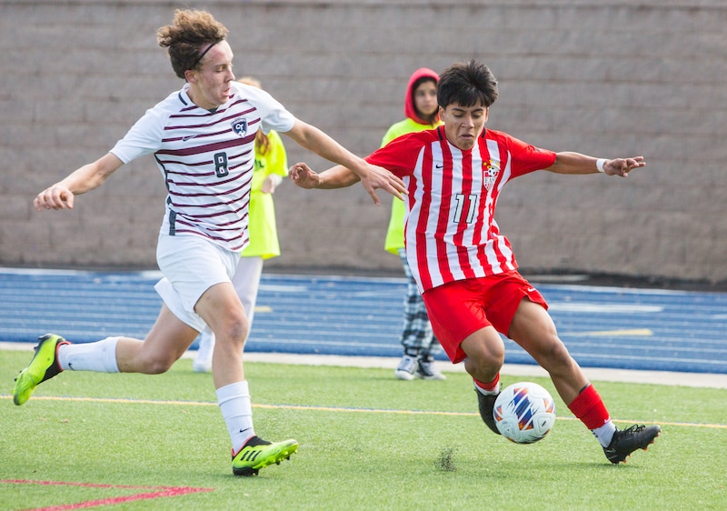 A boy in a red and white soccer uniform plays against a boy in a white and burgundy uniform