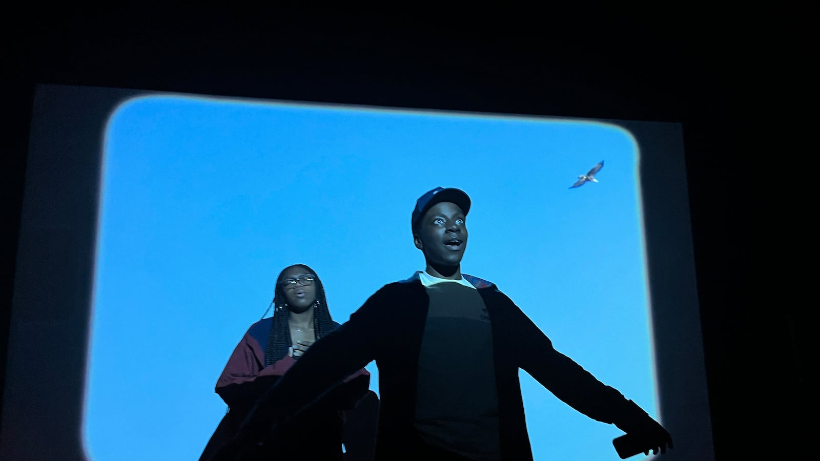 Two high school students stand in the middle of a blue square with a projection of a bird flying above them. 