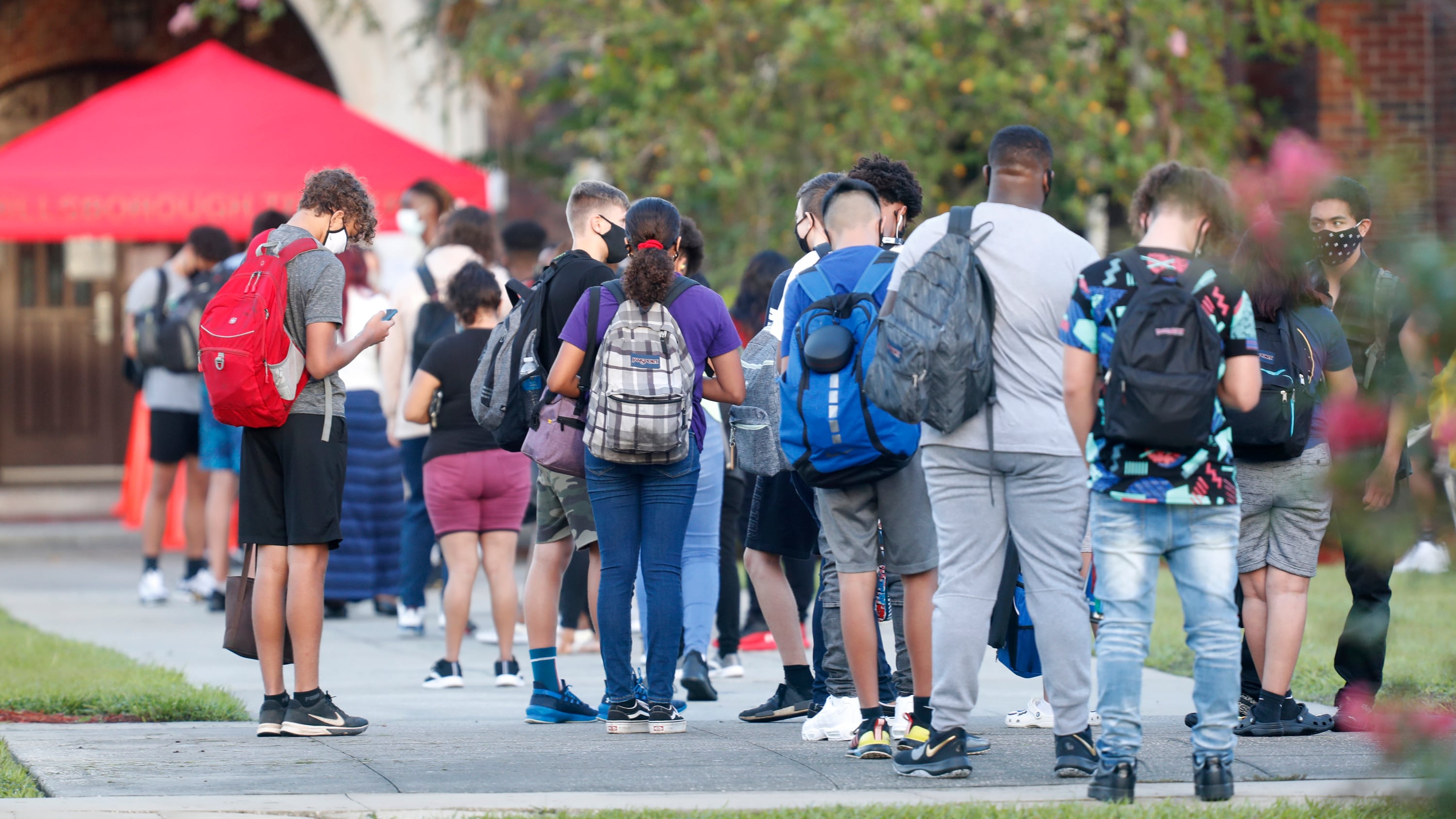 A line of students wearing backpacks and face masks stand in a line outside with trees in the background.