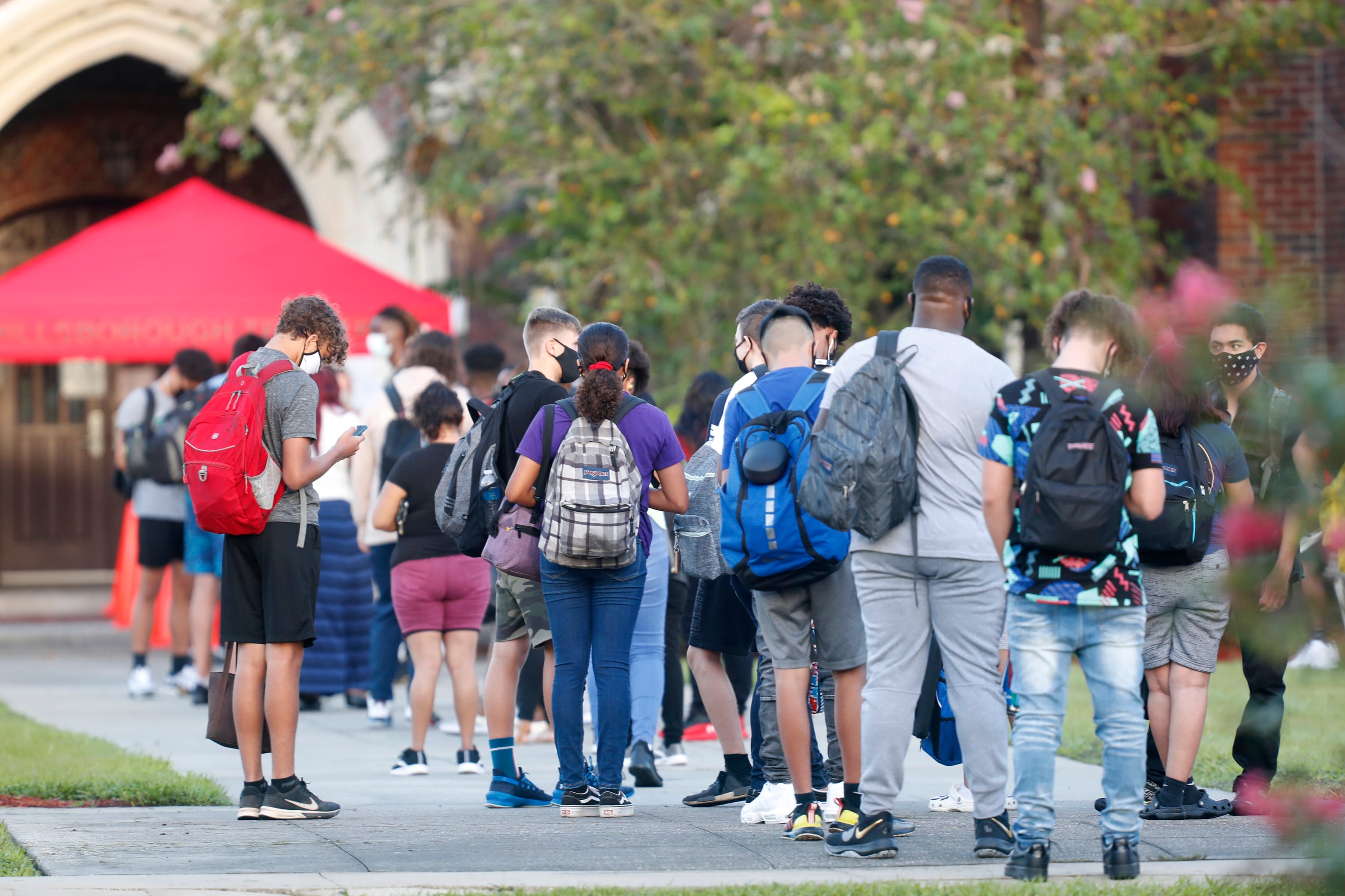 A line of students wearing backpacks and face masks stand in a line outside with trees in the background.
