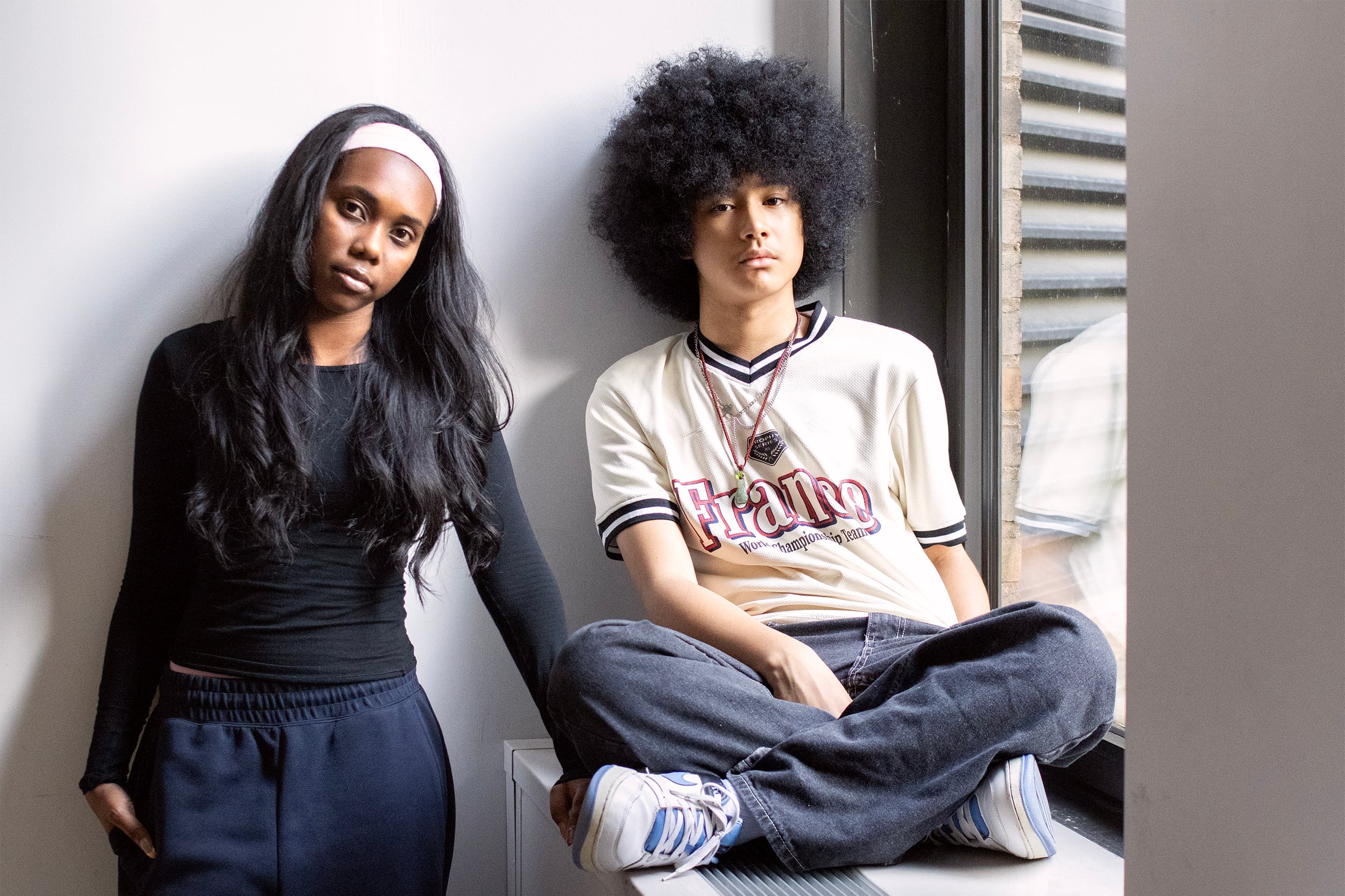 A girl with a white headband stands next to a boy in a white T-shirt seated on a windowsill.