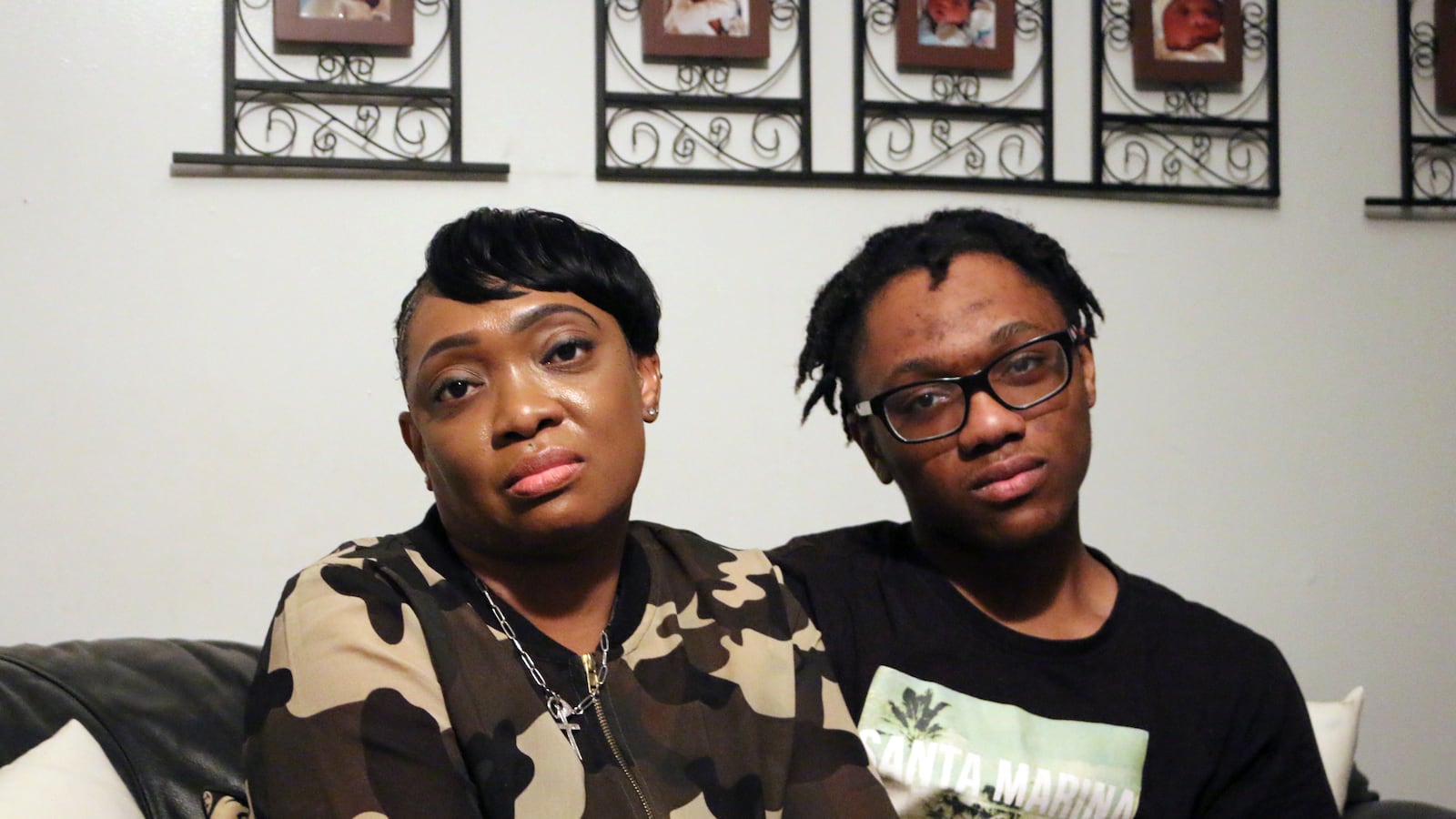A Black mother and son pose for portraits side by side in their home. The teenaged son is wearing glasses and the mom is wearing camouflage sweater. The wall in the background has family photos hanging.