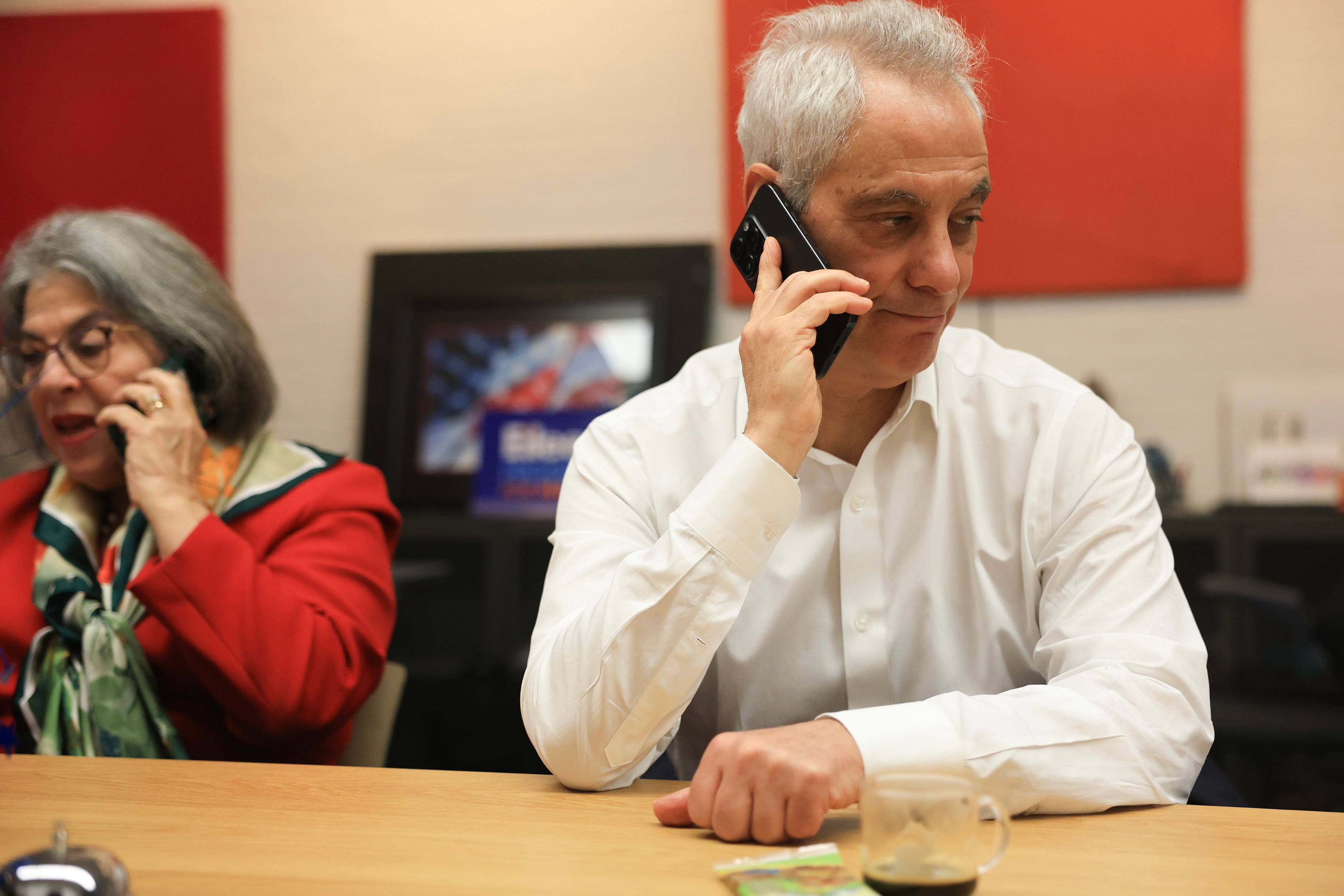 A photograph of a man in a white dress shirt holding a phone to his ear while sitting next to a woman at a wooden table.