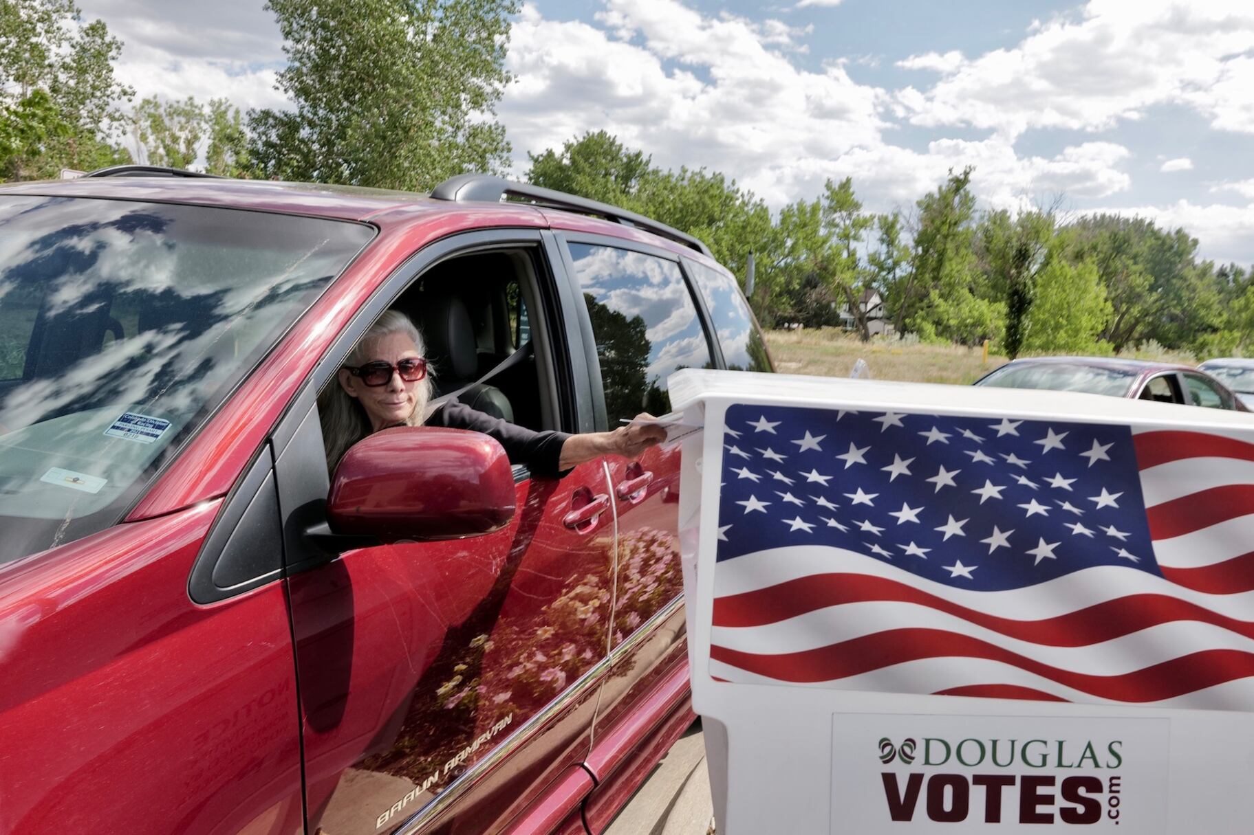 A woman in a red car reaches out of her window toward a box with an American flag.