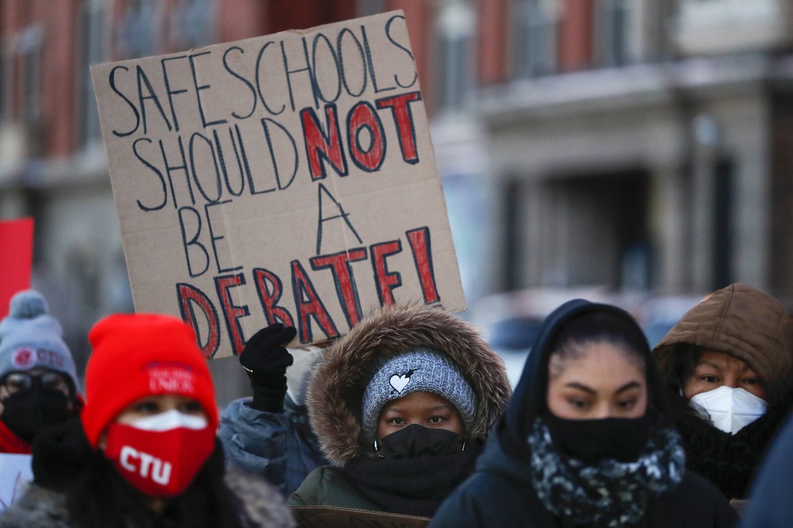 A group of people wearing cold weather clothing stand outside while one person holds up a cardboard sign that reads "safe schools should not be a debate."