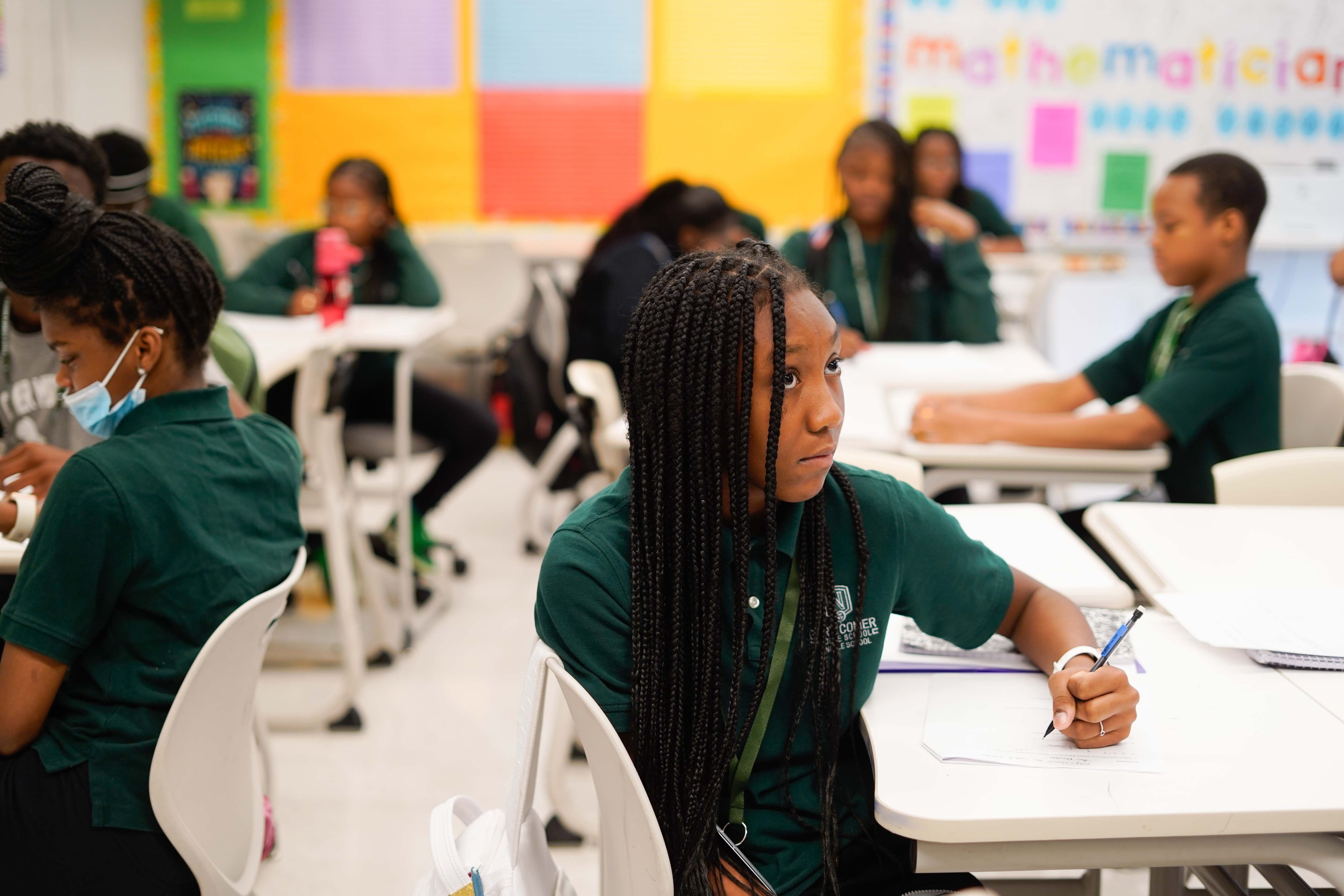 A student with long hair and a green shirt looks up from their desk while completing school work with other students sitting in desks in the background.