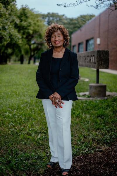 A Black woman in a black blouse stands in a green lawn next to a school building posing for a photograph.