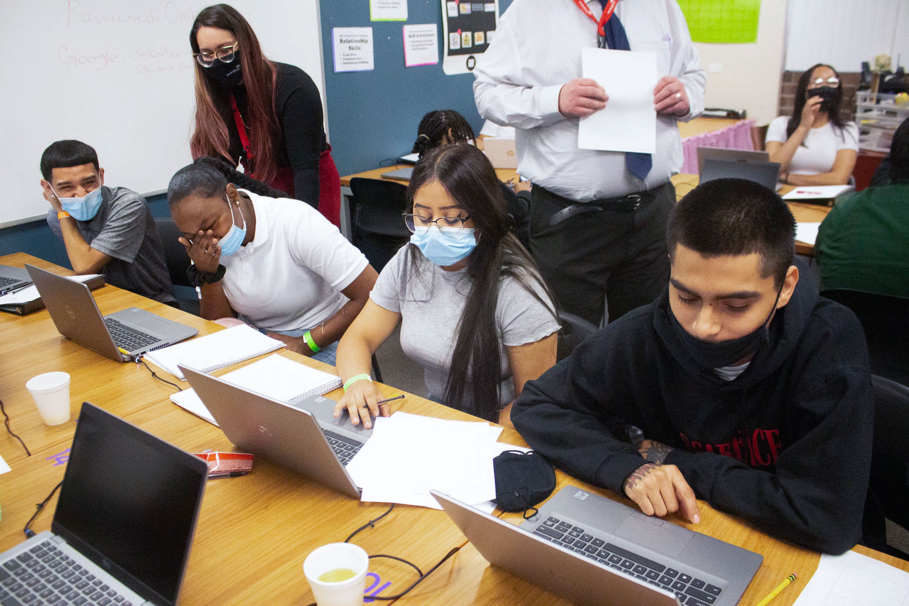 Students work on computers together as teachers look over their shoulders.