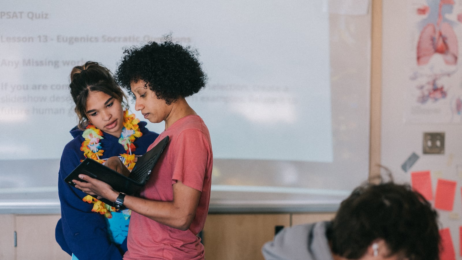 A teacher works with a student at the front of her classroom as another student looks down at his desk in the foreground.