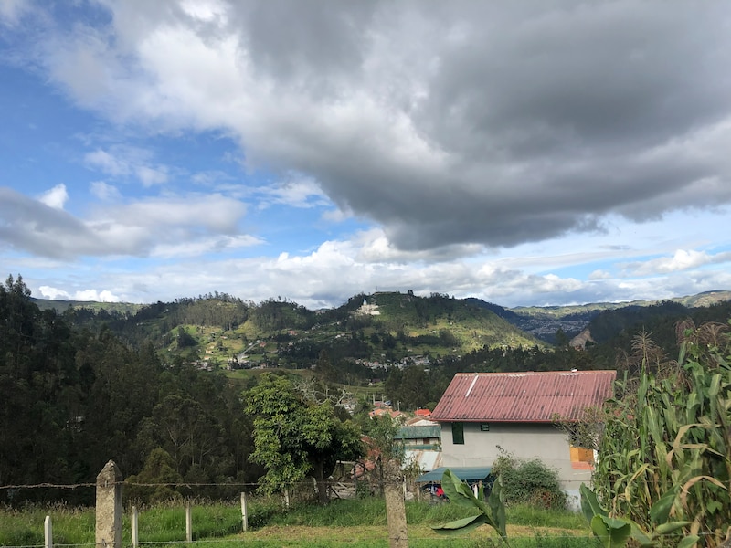 A landscape with a house in the foreground and lush green mountains and a blue sky and clouds in the background.