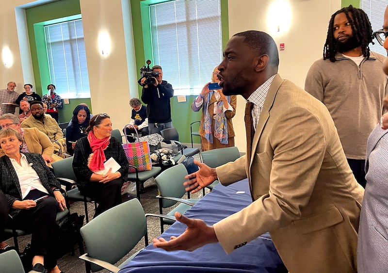A photograph of a Black man in a light colored suit standing in a large meeting room speaking passionately while a group of people stand around him.