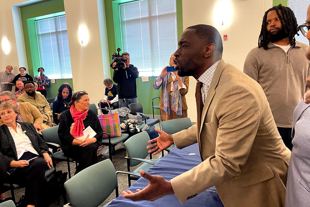 A photograph of a Black man in a light colored suit standing in a large meeting room speaking passionately while a group of people stand around him.