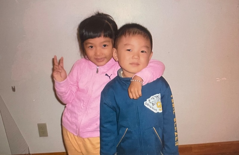 A young girl wearing a pink jacket puts her arm around a young boy wearing a blue jacket.