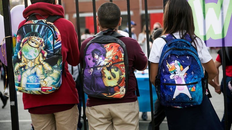 Three students wearing backpacks look through a fence. Their faces aren’t visible.