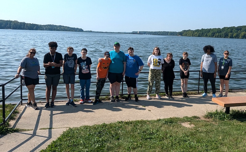 A group of students and a teacher stand on the edge of a body of water in the sun.