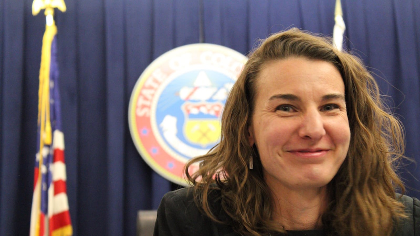 Katy Anthes smiles in front of a state of Colorado seal and an American flag. (photo by Nic Garcia).