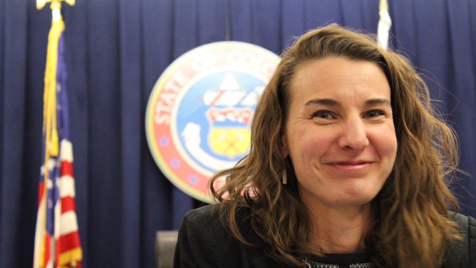 Katy Anthes smiles in front of a state of Colorado seal and an American flag. (photo by Nic Garcia).