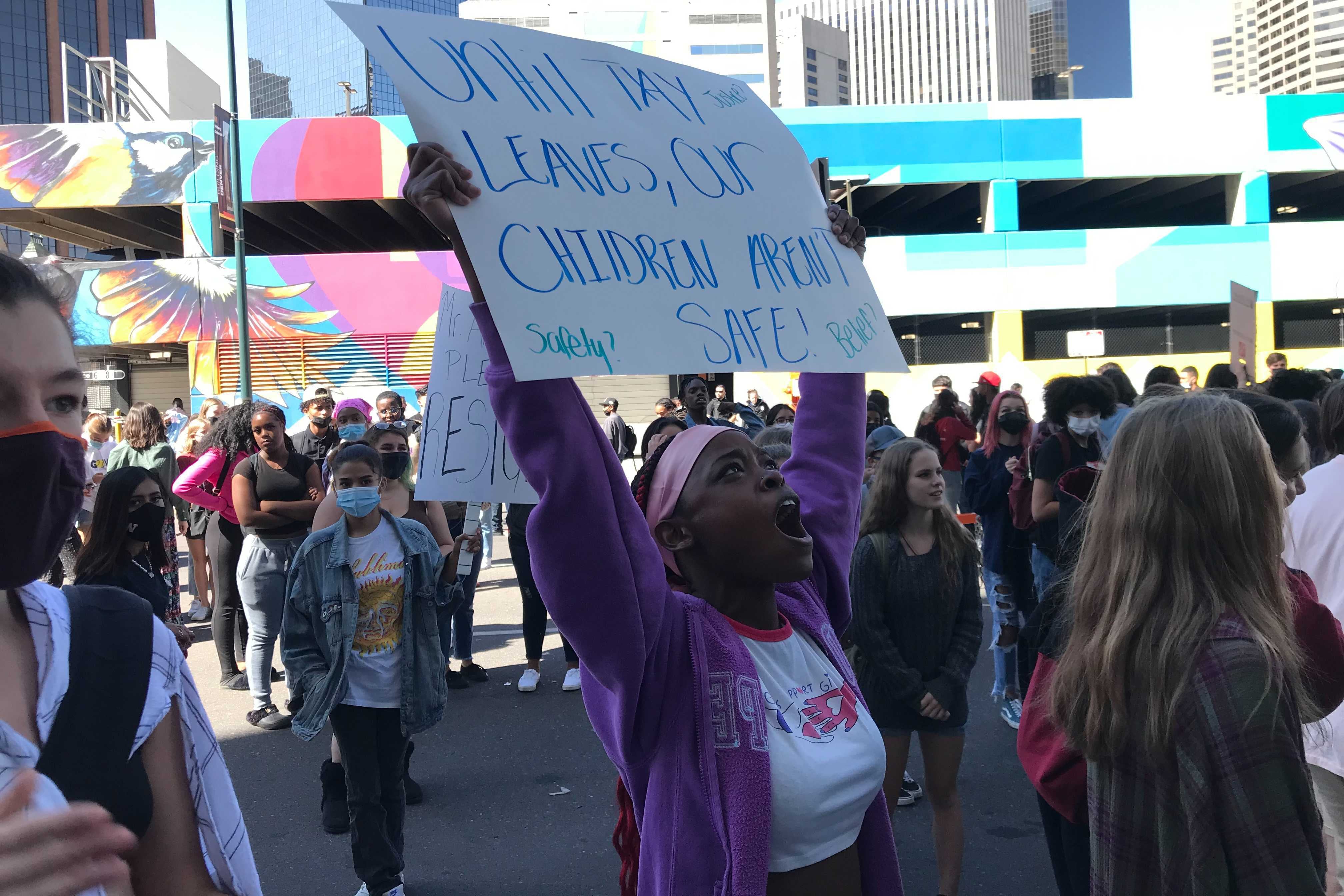 A high school student wearing a purple sweatshirt stands in a crowd. Her arms are raised, holding a sign that reads “Until Tay leaves, our children aren’t safe.” She is shouting.