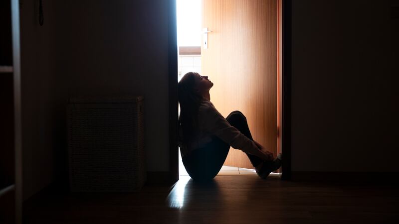 A teenage girl sits in a door frame, silhouetted by light coming in through the door into a dark hallway behind her.
