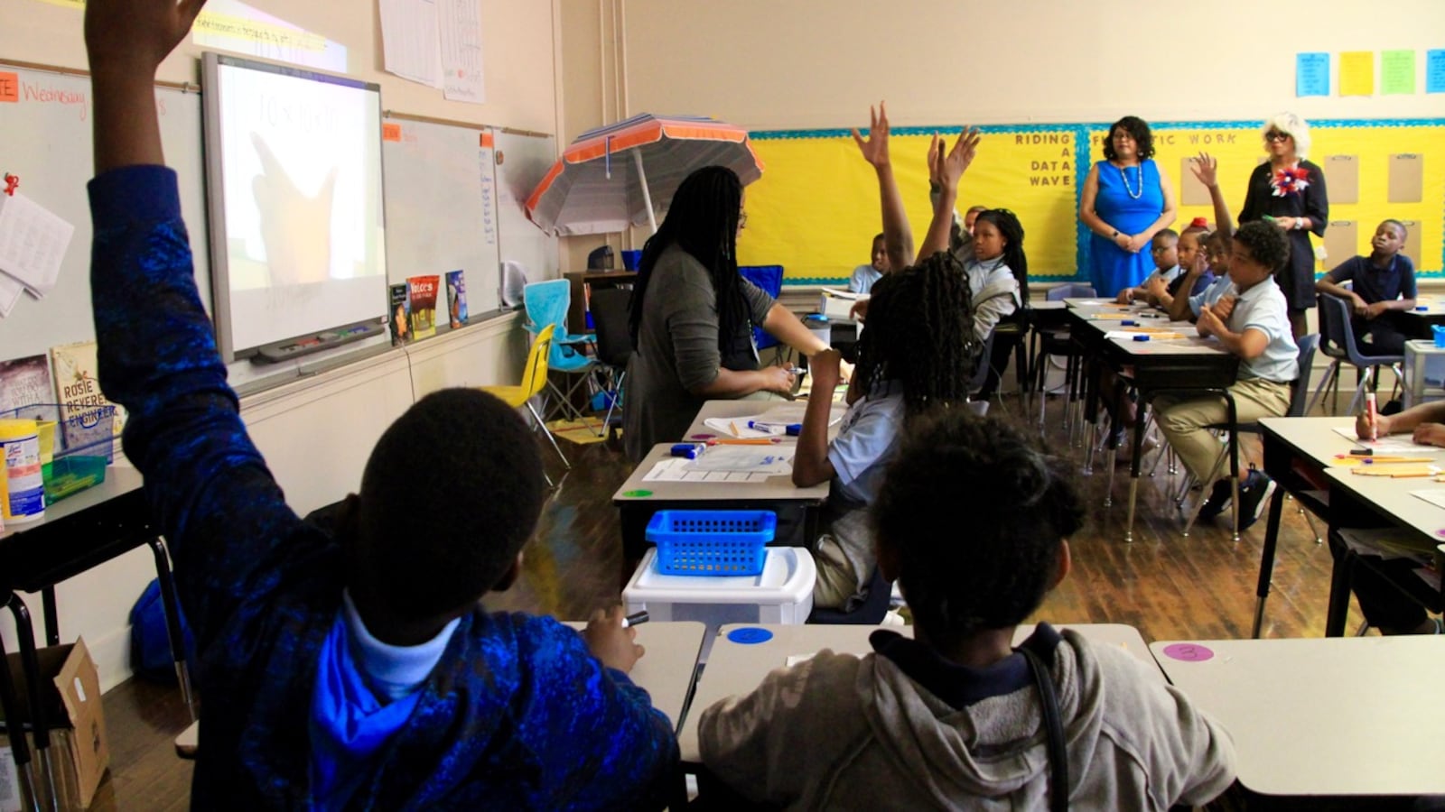 Students and adults sit at desks in a classroom.