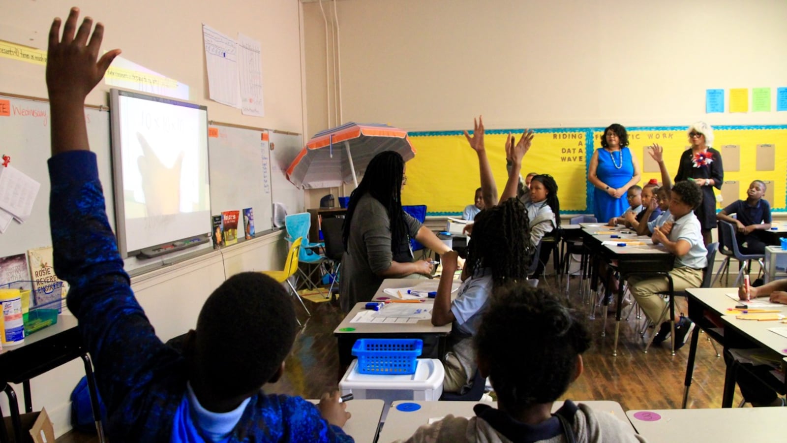 Students and adults sit at desks in a classroom.