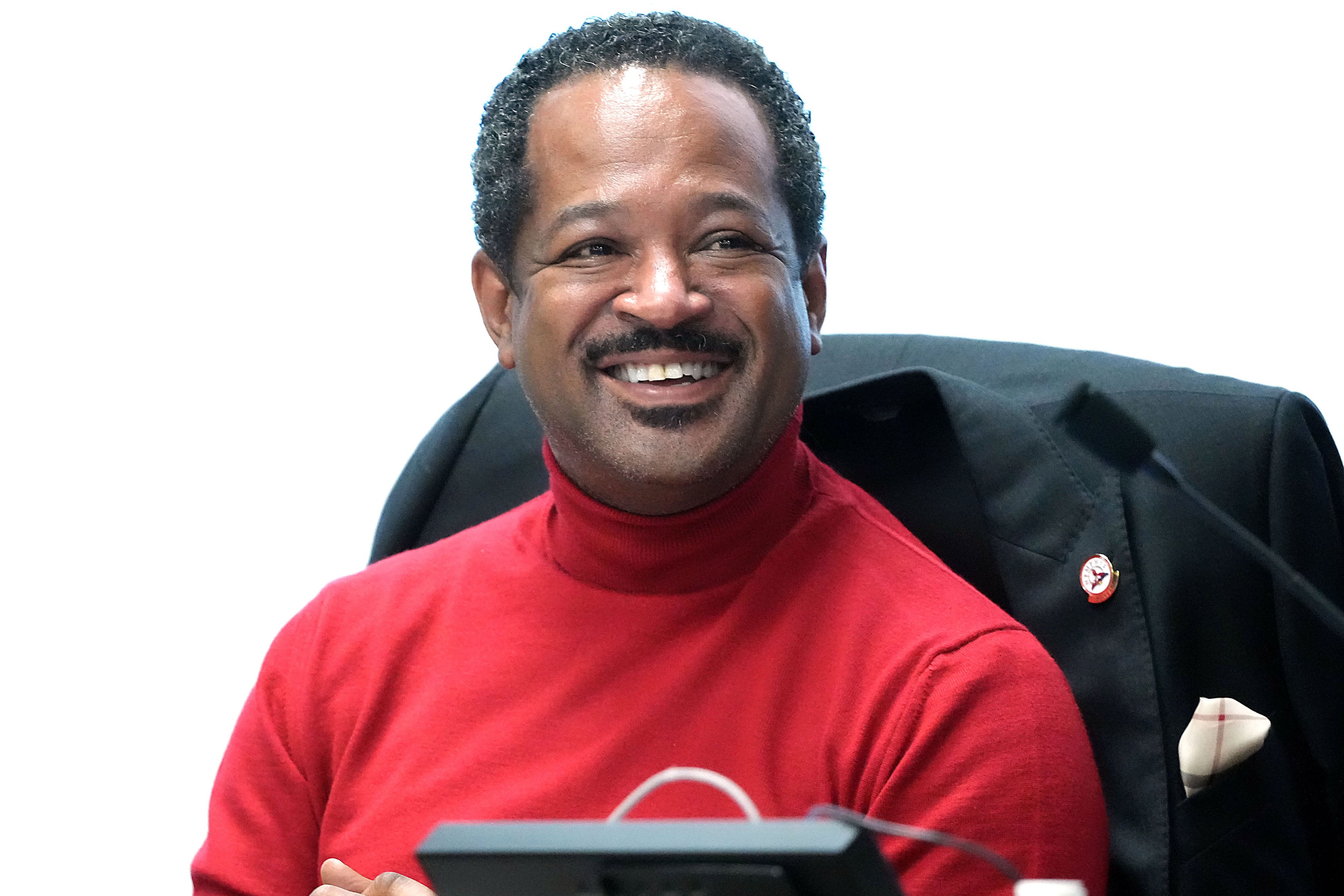 A photograph of a Black man with short dark hair and wearing a red turtleneck sitting at a desk with a big smile on his face.