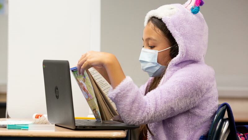 Student wearing a mask and lavender hooded sweatshirt with ears is reading a book in class.