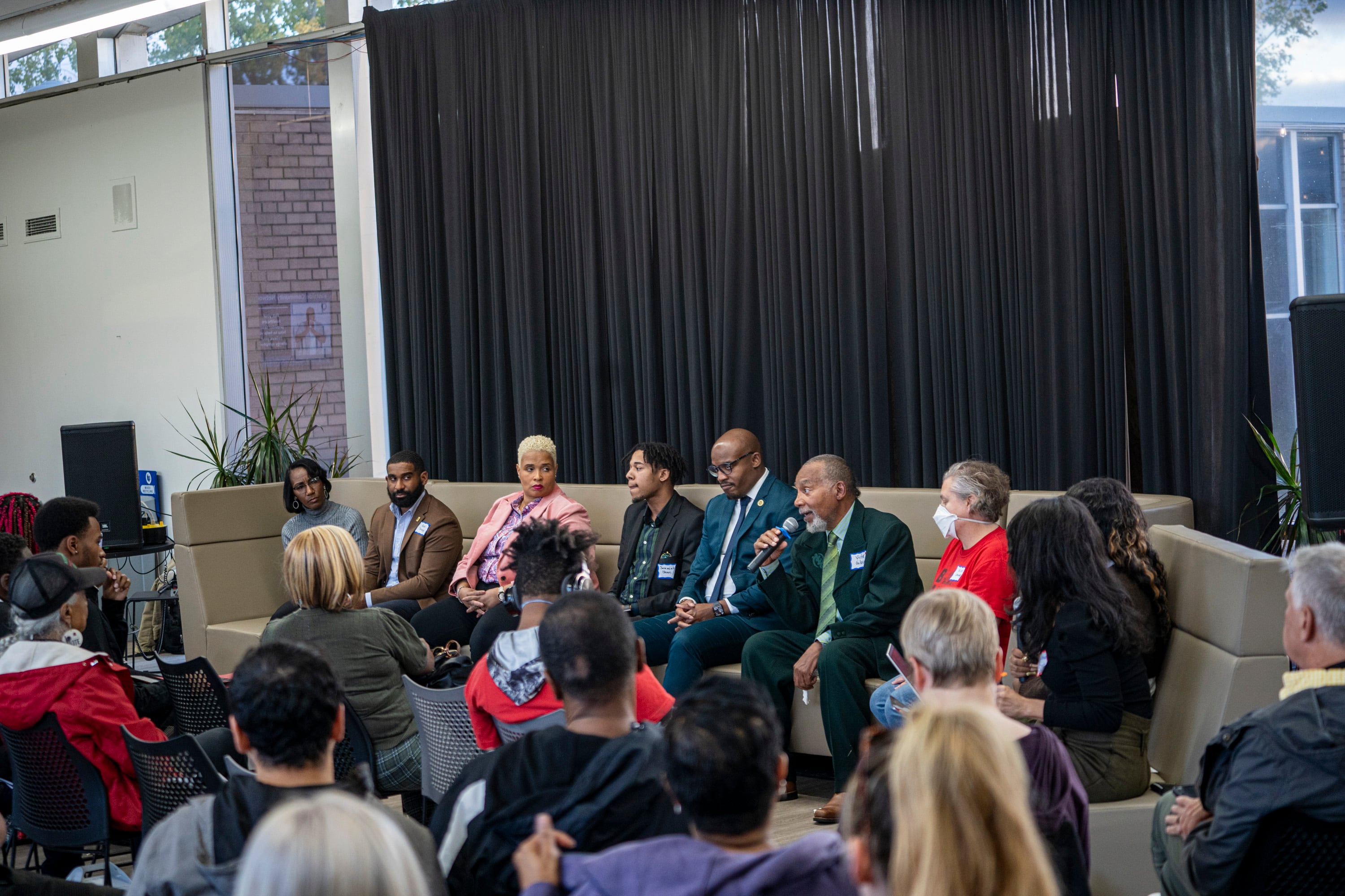 A group of eight people sit on couches, facing an audience of people listening to them speak.