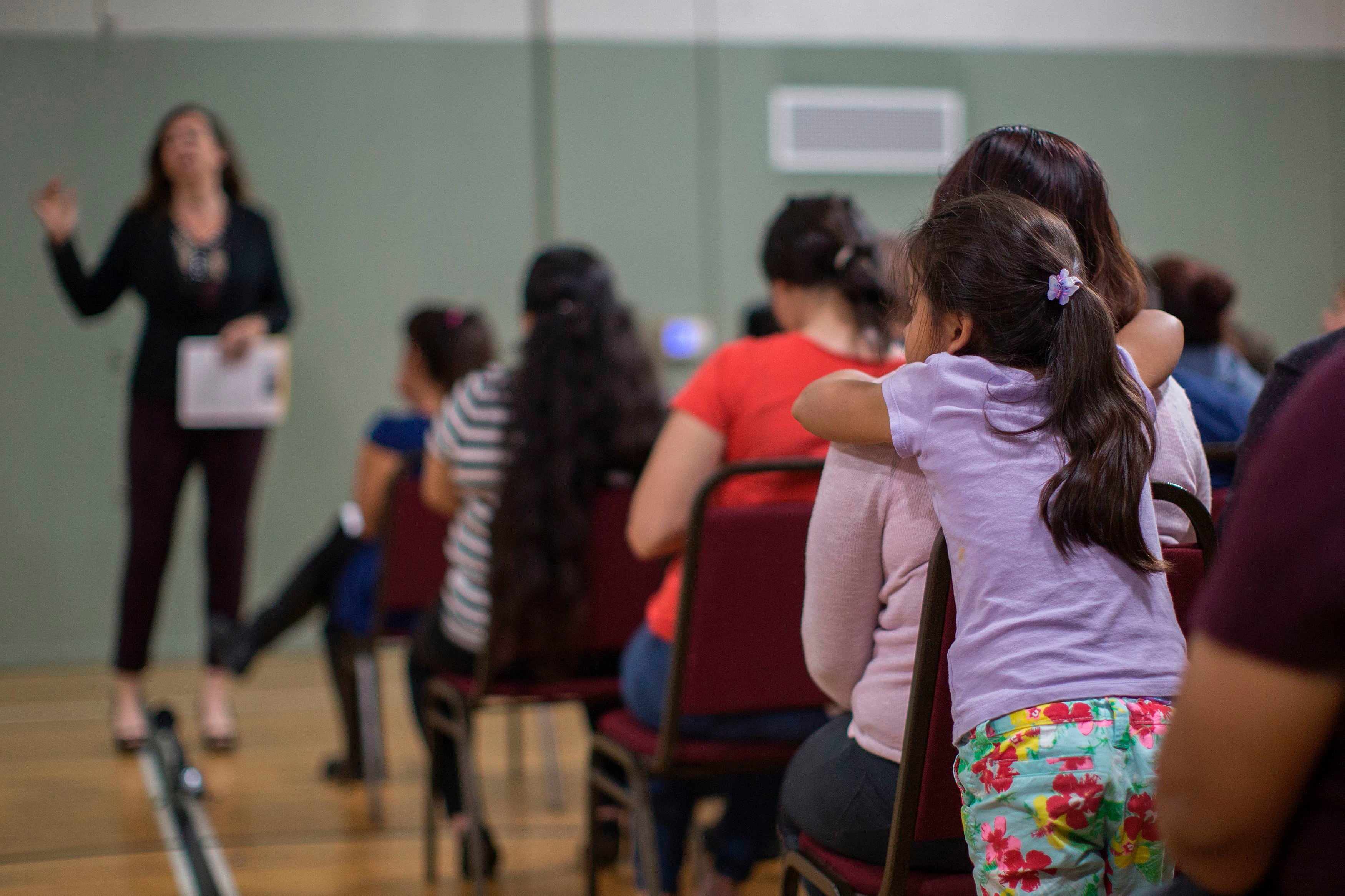 A room full of people sitting in rows of chairs while an adult stands in the background and a young child stands near a chair in the foreground.