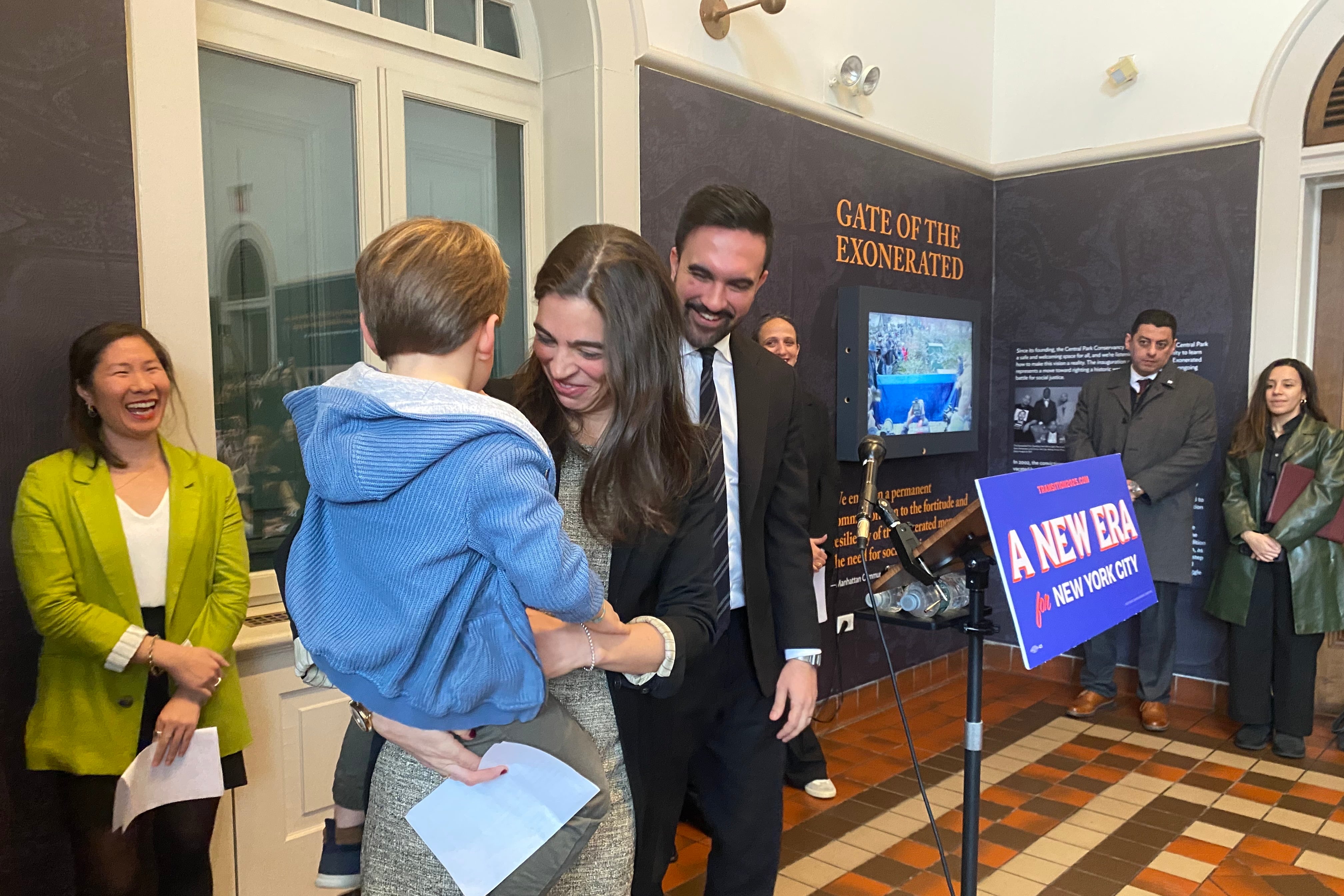 A woman picks up a young boy as a man in a suit smiles.