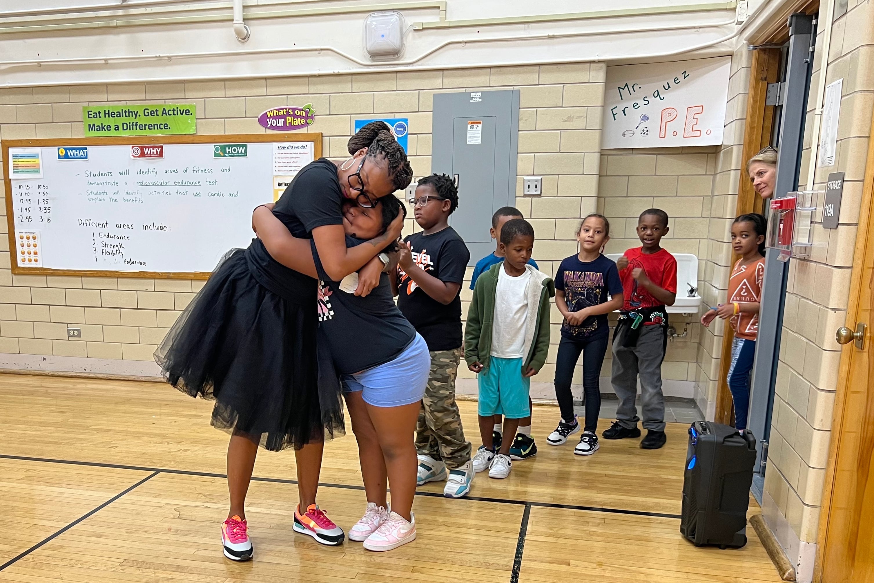 An elementary school principal wearing a tutu skirt hugs a student in the gymnasium.